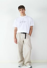 Witte T-shirt met grafische afdruk, beige elastische broek met zwarte riem en grijze sneakers. Eenvoudig ontwerp, relaxed fit.