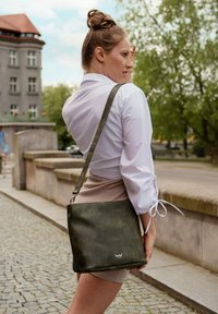 Zelená kožená kabelka přes rameno s hladkým povrchem, čtvercovým tvarem, nastavitelným popruhem a bílým logem na přední straně.