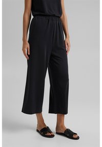Pantalon noir à jambes larges avec une coupe décontractée, doté d'une taille élastique. Porté avec des sandales plates noires, sur un fond neutre.