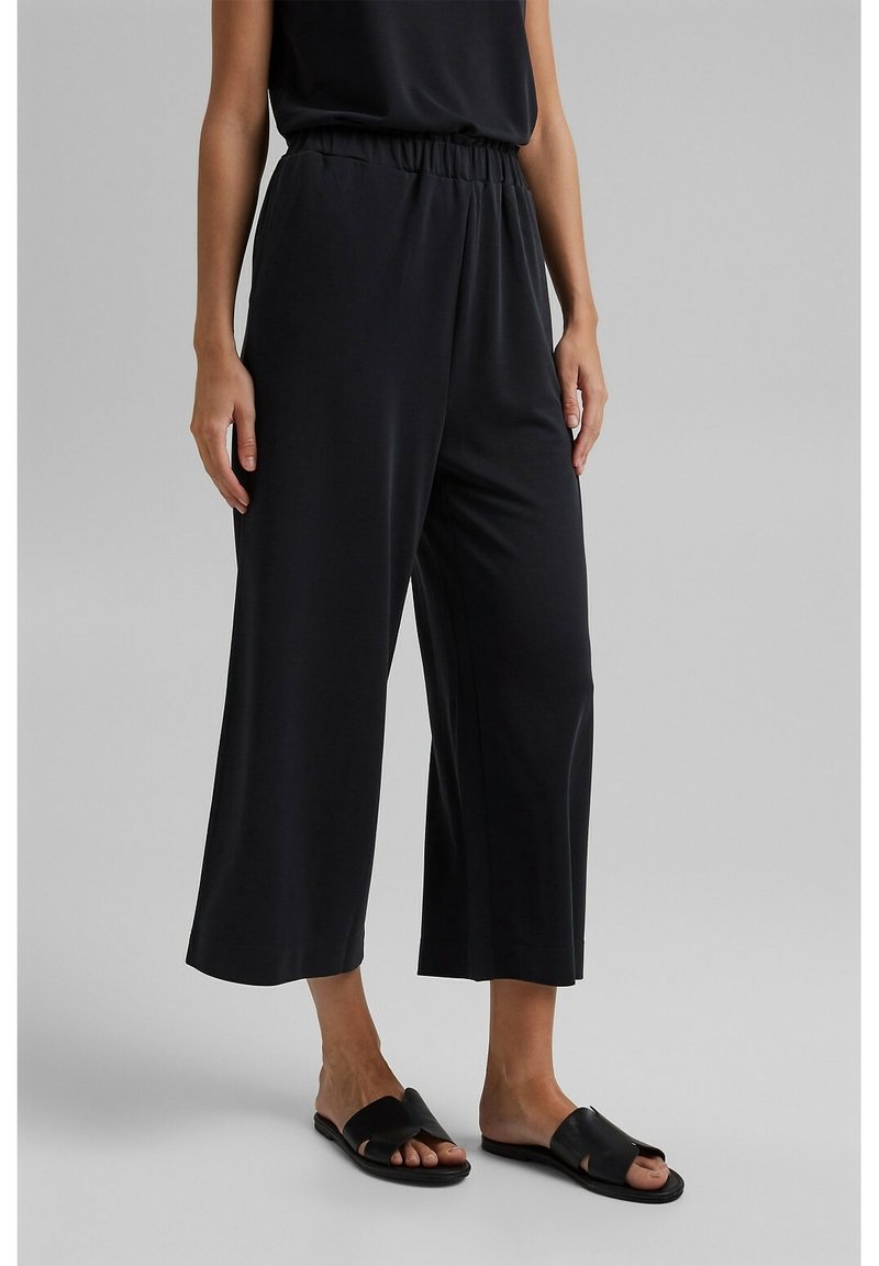 Pantalon noir à jambes larges avec une coupe décontractée, doté d'une taille élastique. Porté avec des sandales plates noires, sur un fond neutre.