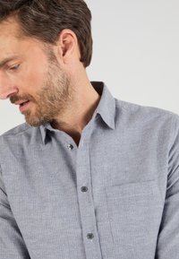 Camicia a maniche corte blu-grigia con texture a spina di pesce, taschino sul petto e bottoni grigi lungo la patta.