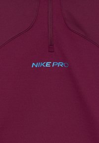 Bordó Nike Pro sportfelső negyedcipzárral és kék fényvisszaverő "NIKE PRO" logóval a mellkason.