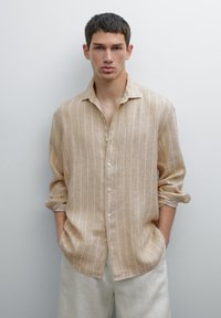 Beige linen shirt with vertical white stripes, long sleeves rolled, button-down front, and relaxed fit. Paired with light-colored trousers.