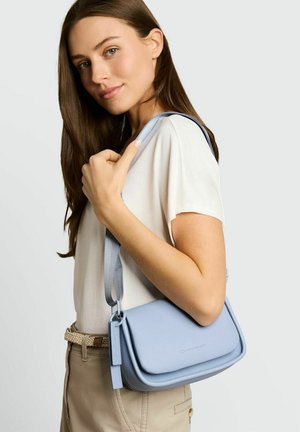 Vrouw met lang bruin haar, gekleed in een wit topje en beige broek, houdt een lichtblauw schoudertasje met een brede band vast.