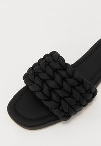 Crna klizna sandal s širokim, pletenim kožnim remenom i ravnim đonom s minimalnim šavovima. Glatka tekstura ukupno, moderan dizajn.