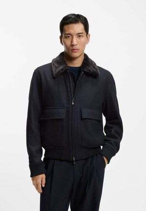 Mężczyzna w ciemnej kurtce zapinanej na zamek, z dwoma przednimi kieszeniami na patki i miękkim kołnierzem, stojący na jednolitym białym tle.