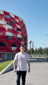 Edifício com padrões geométricos em vermelho e preto, com um exterior brilhante e texturado. Pavimento cinza e vegetação em primeiro plano. Céu azul acima.