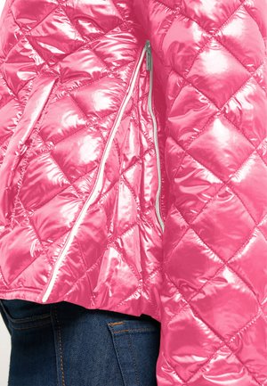 Błyszcząca różowa pikowana kurtka z ukośnym przeszyciem w formie diamentów i pionową srebrną kieszenią na zamek, noszona na ciemnoniebieskich dżinsach.