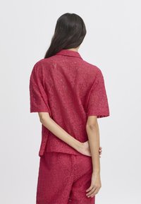 Blusa rossa in pizzo con maniche corte, caratterizzata da un colletto e un motivo floreale. La texture è delicata, con un design a tessitura aperta.