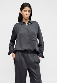 Σκούρο γκρι πουκάμισο με κουμπιά από απαλό ύφασμα με ανάγλυφο σχέδιο και μία τσέπη στο στήθος. Συνδυάζεται με παντελόνι χαλαρής εφαρμογής στο ίδιο χρώμα.