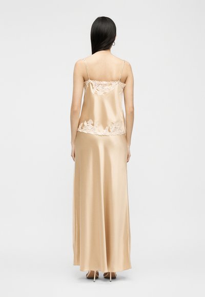 Γυναίκα με μακριά μαύρα μαλλιά που φοράει αμάνικο, μπεζ σατέν τοπ και μακριά φούστα σετ με λεπτομέρειες από λευκή δαντέλα με λουλούδια, στέκεται με την πλάτη προς τα έξω.