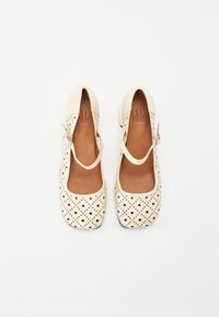 Pantofi din piele de culoare crem, cu un design perforat, vârful rotund și curea la gleznă. Talpă plată și branț maro.