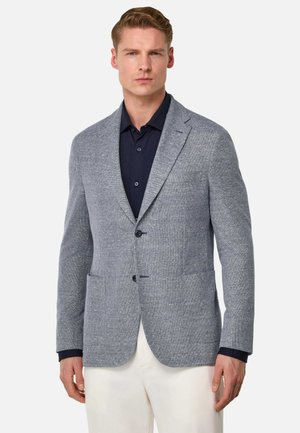 Hellblau strukturierter Blazer mit zwei Fronttaschen, Revers und zwei Knöpfen. Getragen über einem dunklen Hemd; kombiniert mit hellen Hosen.