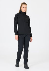 Veste softshell noire avec fermeture éclair à l'avant et poches latérales, associée à un pantalon noir assorti et des chaussures de randonnée sombres. Texture lisse, design ajusté.