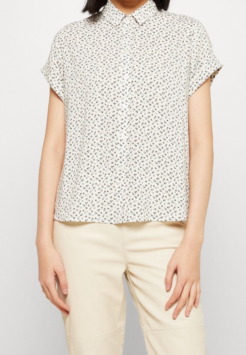 Femme portant un chemisier blanc à manches courtes avec un petit motif croix noir, associé à un pantalon beige taille haute.