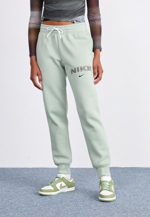 Osoba nosząca jasne zielone spodnie dresowe Nike z białym sznurkiem oraz zielono-białe buty sportowe Nike na szarym dywanie.