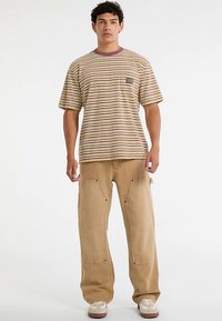 Gestreiftes T-Shirt mit kurzen Ärmeln in Beige mit lila und braunen Streifen, kombiniert mit lockeren braunen Cargohosen mit Seitentaschen und sichtbaren Nähten.