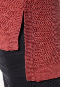 Teksturowany, rdzawego koloru sweter z zygzakowatym wzorem, z bocznymi rozcięciami i ribbowym wykończeniem. Materiał wygląda na miękki i dziany.