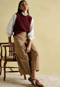Burgunderfarbener Strickpullover über einer weißen Bluse, kombiniert mit braunen karierten weiten Hosen und grünen flachen Schuhen, sitzend auf einem Holzstuhl.