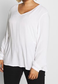 White long-sleeve blouse with a V-neck, lightweight fabric, gathered cuffs, and a relaxed fit, complementing black pants.