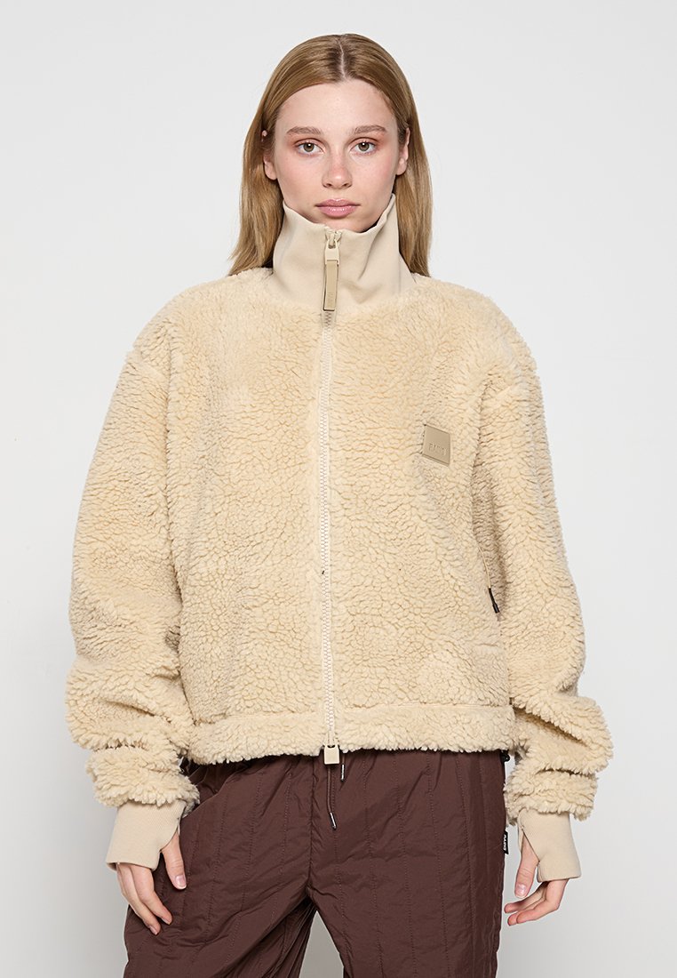 Beige fleecejacka med hög krage, dragkedja fram och elastiska ärmslut. Har en texturerad, fluffig tyg och en liten logotyp patch.