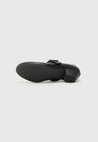 Black shoe with a rubber sole, curved heel, ornamental strap, and detailed tread pattern. Smooth texture and reinforced edges.