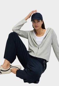 Grau zipfelbare Kapuzenjacke mit geripptem Saum, kombiniert mit dunkelblauen weiten Hosen. Helle Sneakers mit dunklen Streifen. Das Modell trägt eine blaue Kappe.