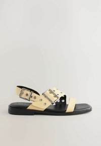 Sandale din piele galbenă cu două curele late, detalii din argint și decupaje circulare. Talpă plată neagră și design cu spatele deschis.