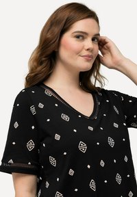 La camiseta negra presenta un escote en V y mangas cortas, decorada con patrones geométricos en blanco y beige. Acento texturizado en el escote.