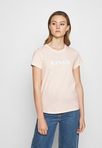 Nuori nainen, jolla on vaaleanruskea hiukset, pukeutuneena vaaleanpunaiseen Levi's t-paitaan ja sinisiin farkkuihin, seisoo tasaisen valkoisen taustan edessä.