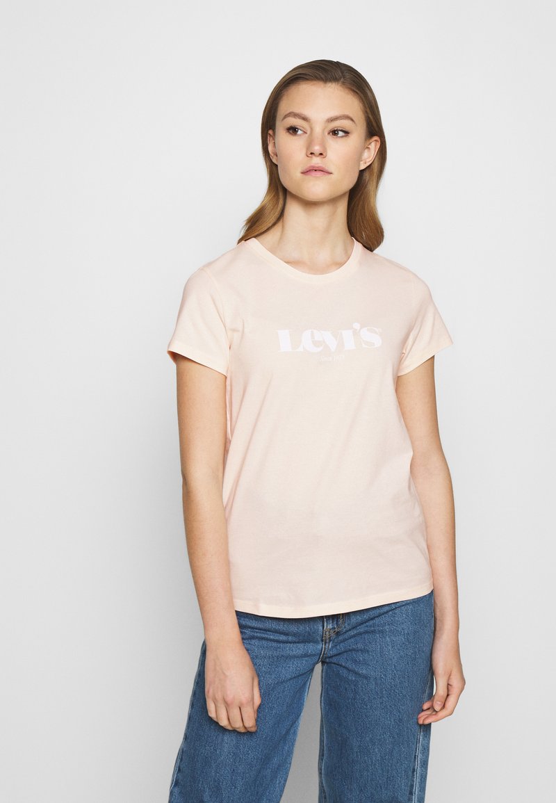 Jonge vrouw met lichtbruin haar, gekleed in een lichtroze Levi's t-shirt en blauwe spijkerbroek, staand tegen een eenvoudige witte achtergrond.