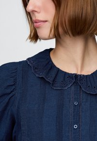 Mörkblå blus med krage prydd med hålmönster, vertikala veck och knappstängning, tillverkad i ett lätt tyg.