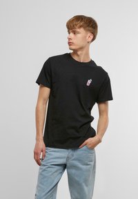 Svart bomull t-skjorte med korte ermer og rund hals, med et lite fargerikt drikkekrusgrafikk på venstre bryst. Kombinert med lyseblå jeans.