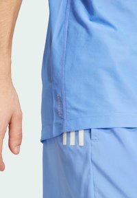 Ljusblå idrottströja med mesh-textur, med en logotyp på sidan och vita ränder på shortsen. Närbild av plaggdetalj.