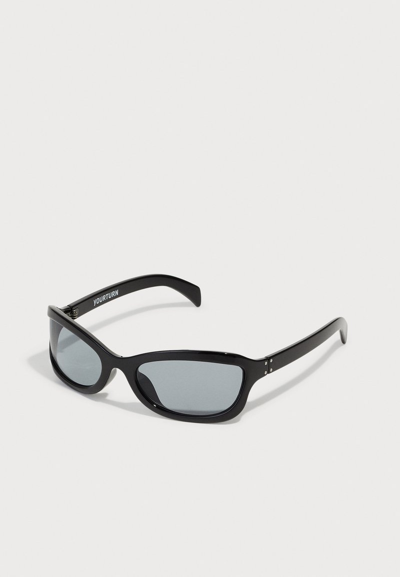 Gafas de sol negras con un diseño angular y elegante. Presentan lentes grises y montura de plástico brillante. Incluyen pequeños acentos metálicos y una marca en la patilla.