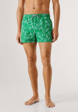 Torso e gambe maschili che indossano pantaloncini da bagno verdi con motivo di foglie bianche, in piedi a piedi nudi contro uno sfondo semplice.