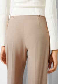 Beige räätälöidyt housut, joissa on sileä pinta, matala vyötärö ja hienovaraiset takasaumadetailit. Yhdistetty valkoisen ribbitun neuleen kanssa.