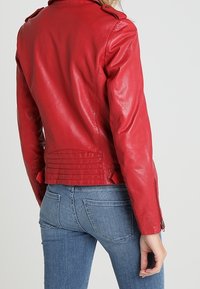 Giubbotto di pelle rossa con una texture liscia, polsini con zip, spalline e pieghe sul retro, abbinato a jeans di denim blu.