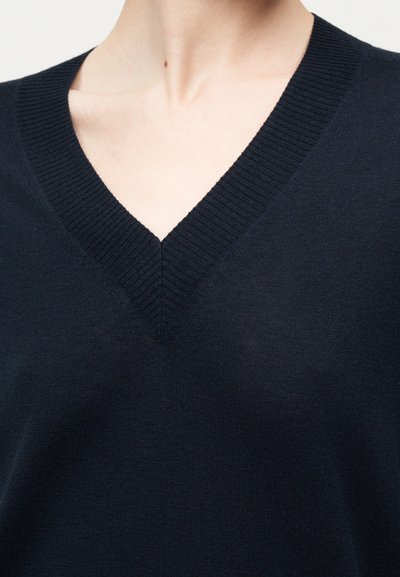 Gros plan d'une personne portant un pull col en V bleu marine foncé avec un col côtelé, montrant le cou et la partie supérieure de la poitrine.