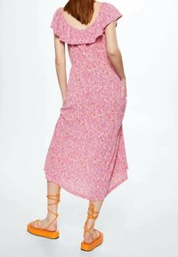 Femme portant une robe midi rose à motifs floraux avec un col à volants et des sandales compensées orange avec des brides à la cheville, debout devant un fond blanc.