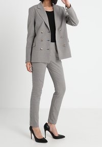 Šedý kostkovaný dvouřadý blejzr se zlatými knoflíky a odpovídajícími slim kalhotami. Černé lodičky na jehlovém podpatku dotvářejí celý outfit.