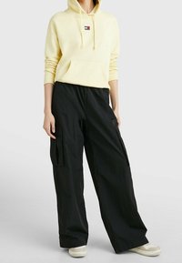 Sudadera amarilla con bolsillo frontal y cordón, combinada con pantalones cargo de pierna ancha en negro, que cuentan con bolsillos laterales y una textura suave.