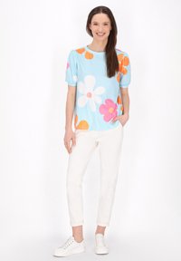 Coprire a maniche corte di colore azzurro chiaro con grandi motivi floreali multicolori in rosa, arancione e bianco; abbinato a pantaloni bianchi arrotolati e sneakers bianche.