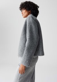 Giacca grigia intrecciata con una finitura testurizzata, dal design cropped e maniche lunghe. Abbinata a pantaloni aderenti grigio chiaro.