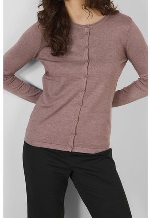 Kvinde med bølget hår iført en mauve knappet cardigan og sorte bukser, stående med hænderne på hofterne mod en ensfarvet baggrund.