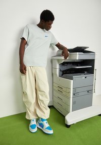 T-shirt Nike gris clair, pantalon ample crème, baskets bleu et gris, appuyé contre une imprimante multifonction sur un tapis vert.