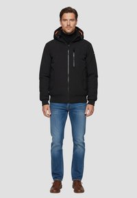 Schwarze Kapuzenjacke mit orangenen Akzenten, Reißverschlusstaschen und strukturierter Oberfläche, kombiniert mit blauen Jeans und braunen Schuhen.