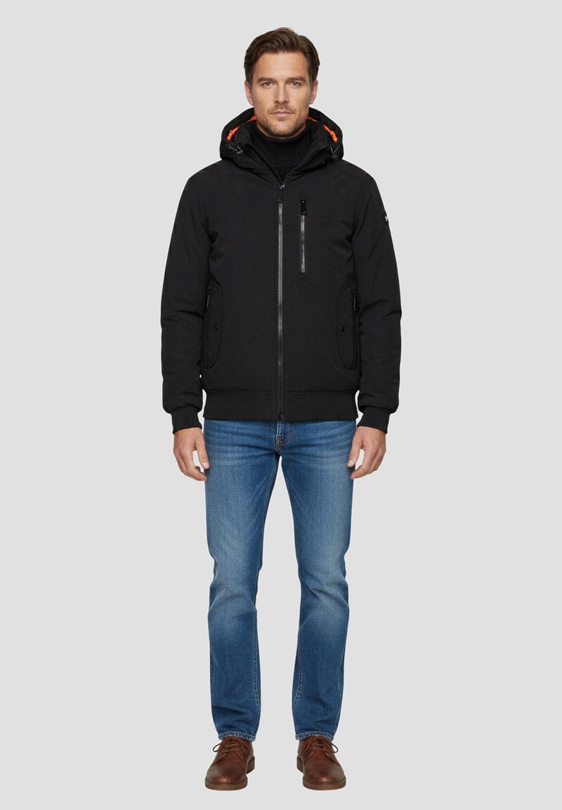 Schwarze Kapuzenjacke mit orangenen Akzenten, Reißverschlusstaschen und strukturierter Oberfläche, kombiniert mit blauen Jeans und braunen Schuhen.