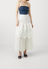 Top sin tirantes de mezclilla combinado con una falda blanca de encaje con volantes en capas y detalles de ojal. Incluye tacones abiertos negros.