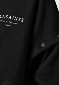 Melns džemperis ar teksturētu virsmu, uz kura ir balts logotips un melnas spraudņa pogas gar plecu šuvi.